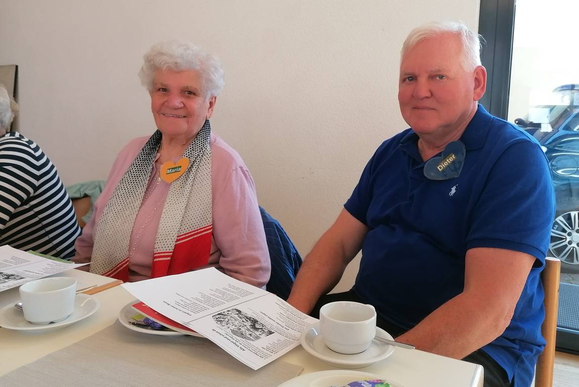 Bild mit Gästen und einem Betreuer an der Kaffeetafel mit Programmheft
